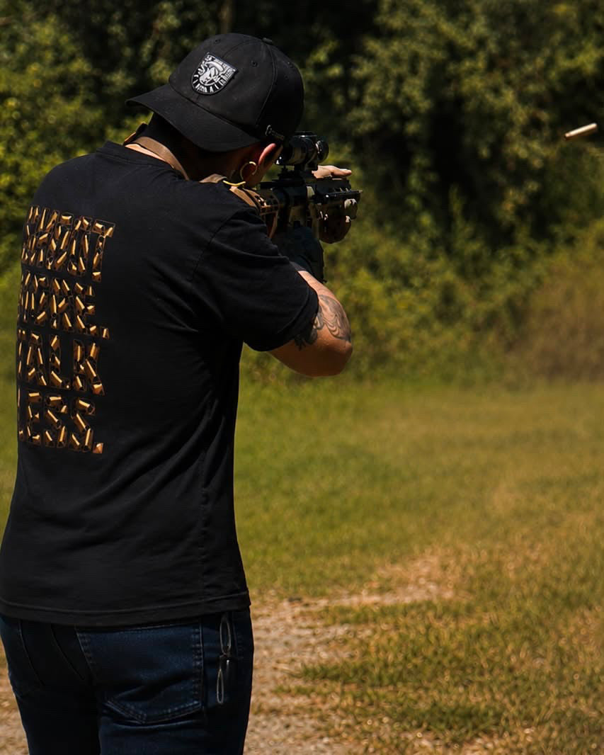 Shoot More Talk Less tee with brass lettering worn at an outdoor range